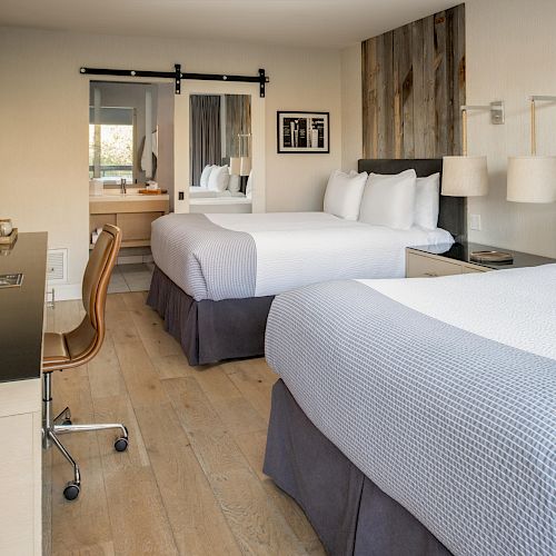 The image shows a hotel room with two beds, a desk with a chair, a mirror, and a bathroom in the background. The decor is modern and cozy.