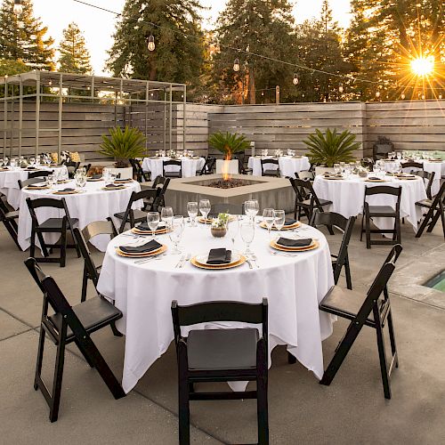 An outdoor dining setup with round tables, black chairs, white tablecloths, and place settings, positioned around a central fire pit and beside a hot tub.