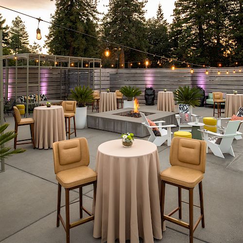 An outdoor event space with tables, chairs, string lights, and potted plants, and a hot tub on a concrete patio. There are trees in the background.