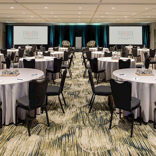 A conference room set up with round tables, chairs, two projection screens, and a stage with chairs and a table, ready for a meeting or event.