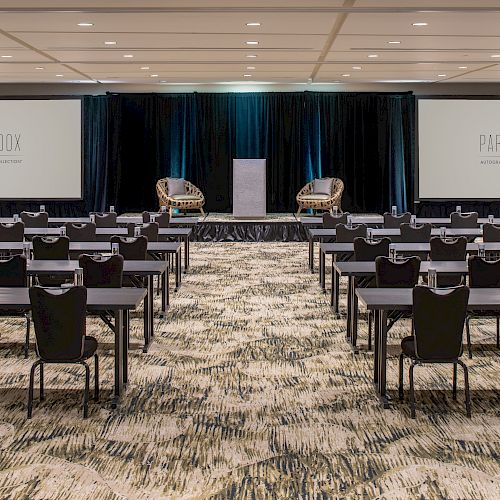 The image depicts a conference room setup with rows of tables and chairs facing a stage with two armchairs. Two screens display 