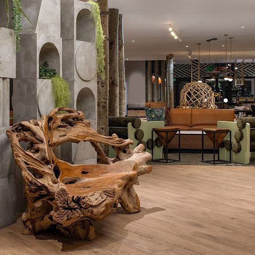 The image shows a stylish interior with a unique wooden bench, modern seating, greenery, and decorative elements in a spacious open area.