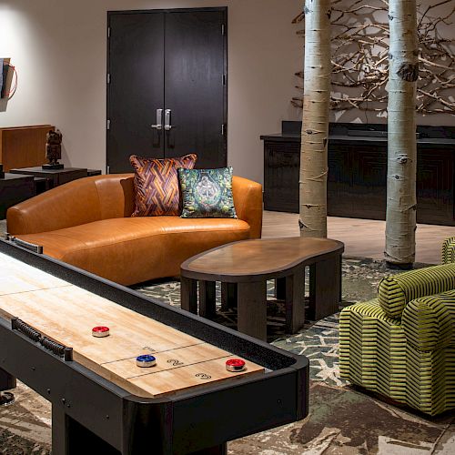 A cozy lounge area features a shuffleboard table, a brown leather couch, vibrant cushions, a wooden coffee table, and tree-like decor elements.