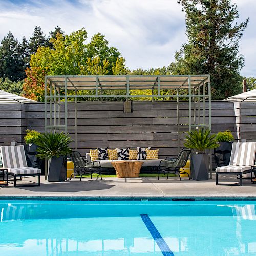 A serene poolside view with lounge chairs, umbrellas, potted plants, and a pergola-covered seating area surrounded by wooden fencing and greenery.