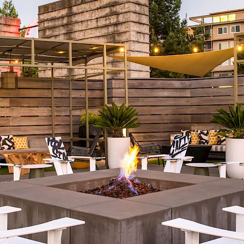 The image shows an outdoor seating area with a fire pit at the center, surrounded by white chairs and potted plants, and string lights above.