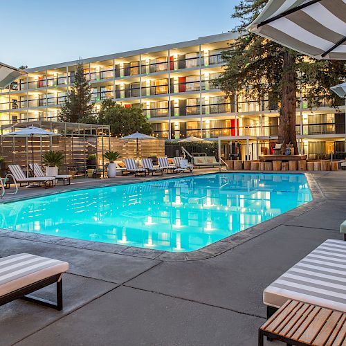 The image shows a well-lit outdoor swimming pool area with lounge chairs and umbrellas, surrounded by a multi-story building with balconies.