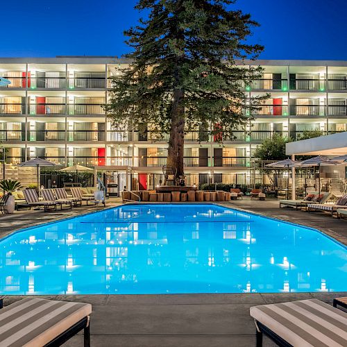 This image shows a brightly lit hotel pool area at night, with lounge chairs, umbrellas, and a multi-story building in the background.