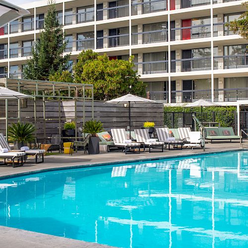 A clear blue swimming pool with lounge chairs, umbrellas, and a modern apartment building in the background.