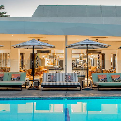 A luxurious poolside area featuring cushioned lounge chairs, umbrellas, and a view into a chic indoor area with modern furnishings.