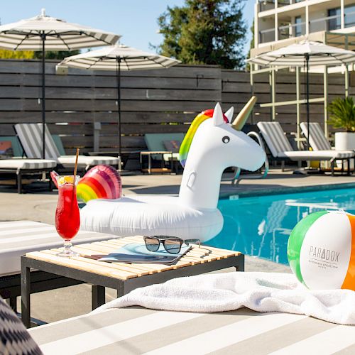 A poolside scene with a unicorn float, cocktail, beach ball, lounge chairs, and umbrellas. A towel is draped on a chair.