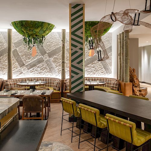 A modern, stylish restaurant with green chandeliers, yellow seating, a long table, and a textured wall featuring abstract artwork.