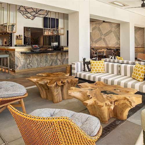 A stylish, modern lounge area with wooden tables, comfy seating, accentuating striped cushions, and vibrant decor, adjacent to a well-lit bar area.