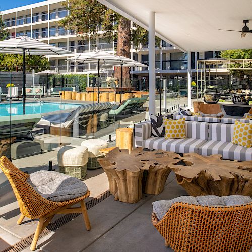 Outdoor lounging space by a pool with modern furniture, wooden tables, striped cushions, and umbrellas in a sunny setting.