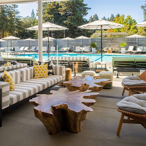 Outdoor seating area with striped couches, wooden tables, and cushioned chairs by a pool with lounge chairs, greenery, and umbrellas in the background.