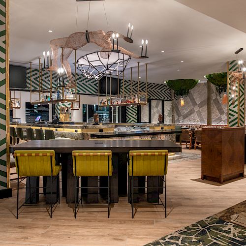 A modern bar with patterned walls, a central bar area with green and yellow seats, hanging plants, and eclectic decor, featuring contemporary lighting.