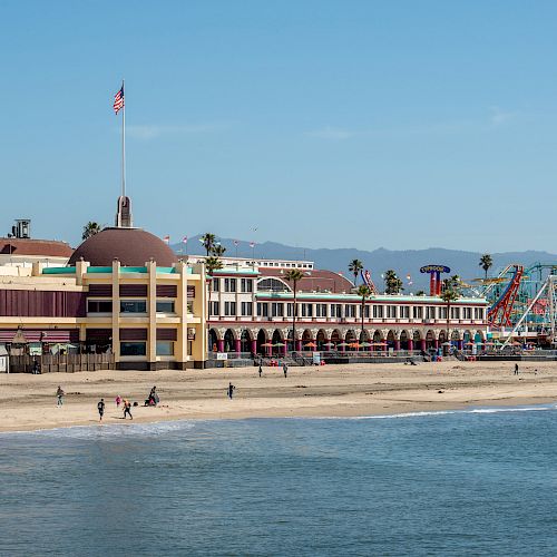 A beachside amusement park with a historic building, a roller coaster, palm trees, and people walking on the sandy beach, under a clear blue sky.