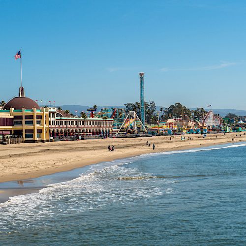 A beachside amusement park with various rides, a building with a dome and flag, calm waters, clear skies, and people walking on the sandy shore.