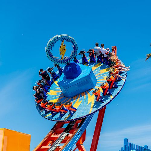 A vibrant amusement park ride with people seated on a rotating and tilting disc, with a clear blue sky and a palm tree in the background.