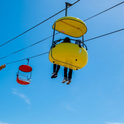 The image shows people riding colorful gondolas on a sky ride against a clear blue sky, with their legs dangling from the colorful pods.