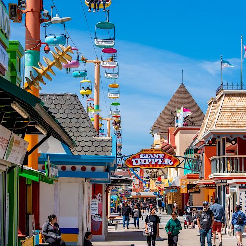 A vibrant, colorful amusement park scene with people walking, shops, and a chairlift ride extending into the distance under a bright blue sky.