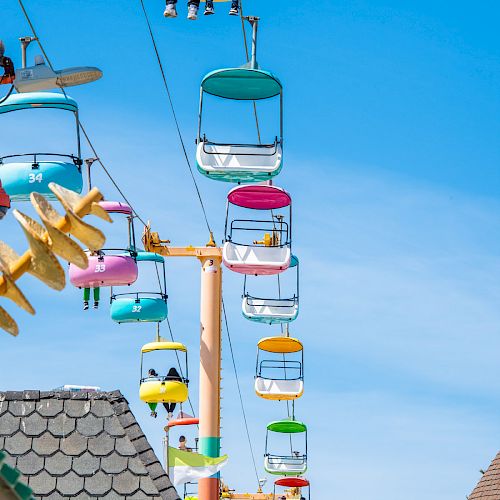 People riding colorful gondolas on a cable lift against a clear blue sky, with rooftops visible at the bottom.