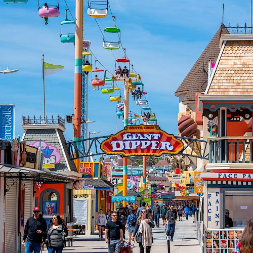 People walk along an amusement park boardwalk featuring colorful sky rides overhead and a prominent sign reading 