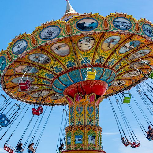 Colorful amusement park swing ride with people seated in chairs, suspended by chains, spinning in the air, set against a bright blue sky.