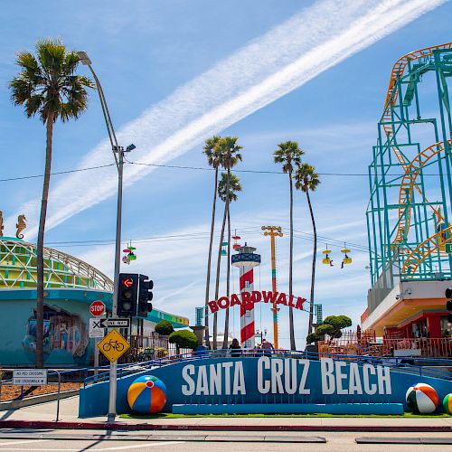 The image shows the entrance to Santa Cruz Beach Boardwalk with palm trees, roller coasters, and signage against a clear blue sky.