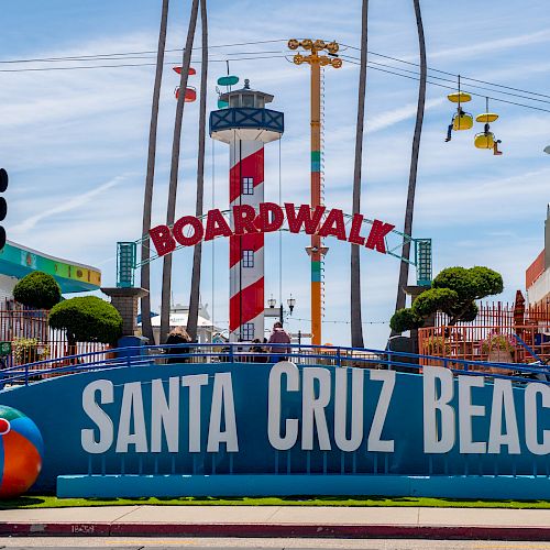 Entrance to Santa Cruz Beach Boardwalk with a lighthouse, colorful signs, and decorations. Sky chairs and a roller coaster are also visible.