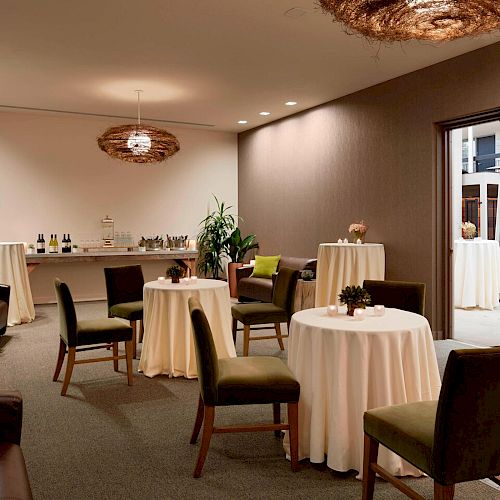 The image shows a cozy room set up for a small gathering, with round tables covered in white tablecloths, chairs, and a refreshment table.