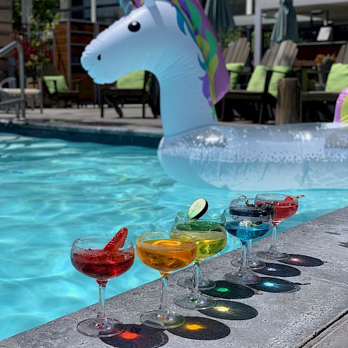 A unicorn pool float and five colorful cocktails lined up by the poolside, with patio chairs and umbrellas in the background.