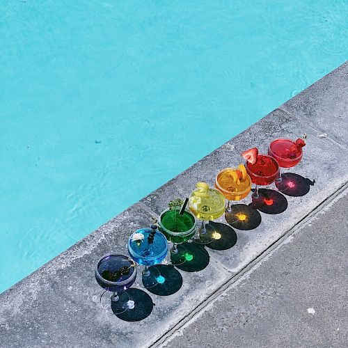 A lineup of colorful drinks, each a different color of the rainbow, placed by the edge of a swimming pool.