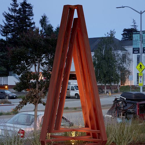 A triangular metal sculpture is set in an urban area with grass and concrete blocks at its base. The moon is visible in the evening sky.