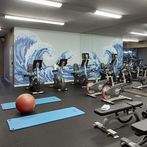 A gym with exercise equipment including bikes, treadmills, weights, and benches, alongside mats and a ball, with a wave mural on the wall.