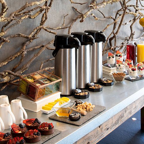 A breakfast buffet with coffee dispensers, yogurt parfaits, juice, pastries, mini cakes, tea bags, and sauce containers on a wooden table.