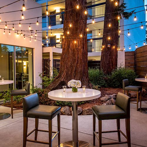 An outdoor patio with string lights, high tables, and chairs, decorated with flower arrangements, set against a backdrop of trees and a modern building.