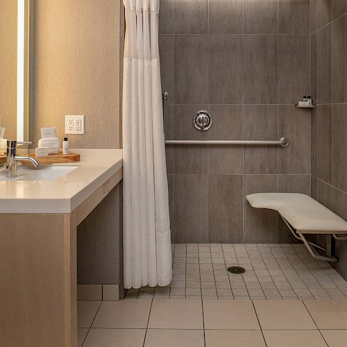 The image shows a modern bathroom with a vanity sink, a well-lit mirror, a walk-in shower with a white curtain, and a foldable shower seat.