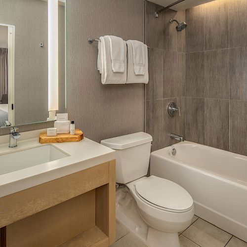 The image shows a modern bathroom with a bathtub, toilet, sink, and a large mirror. Towels are neatly hung above the bathtub.