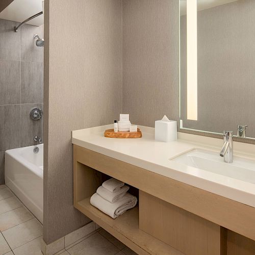 A modern bathroom featuring a shower-bathtub combo, toilet, large mirror with lights, sink, countertop with toiletries, and neatly folded towels below.