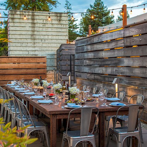 An outdoor dining area with a long wooden table set with plates, glasses, and flowers, surrounded by high wooden fences and string lights.