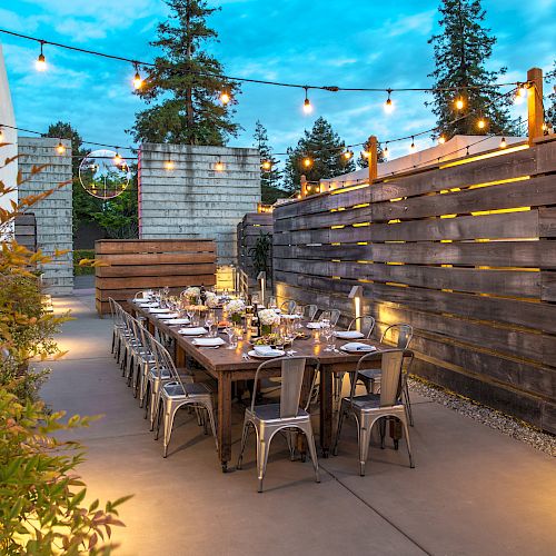 An outdoor dining area features a long wooden table set for a meal, string lights overhead, and surrounded by wooden and plant décor, creating a cozy atmosphere.