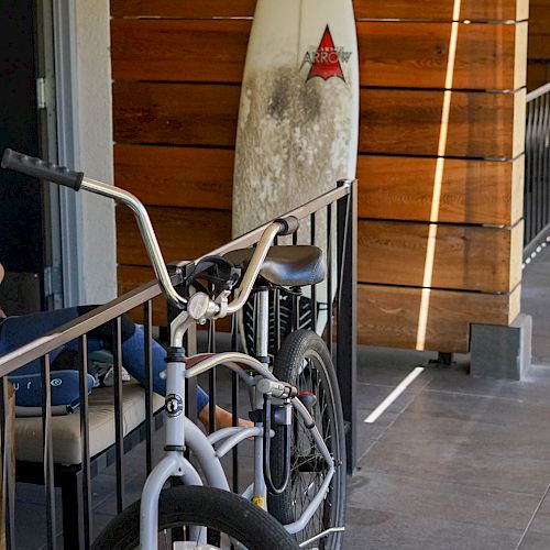 The image shows a bicycle parked next to a balcony railing, with a surfboard standing upright against a wooden divider in the background.