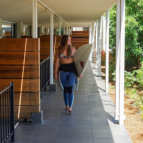 A person walks along a covered walkway, carrying a surfboard under their arm, surrounded by greenery and wooden structures at their side.