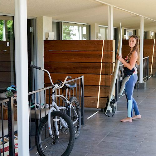 A person in a wetsuit stands with a surfboard beside a parked bicycle in an outdoor area featuring wooden dividers and concrete flooring.