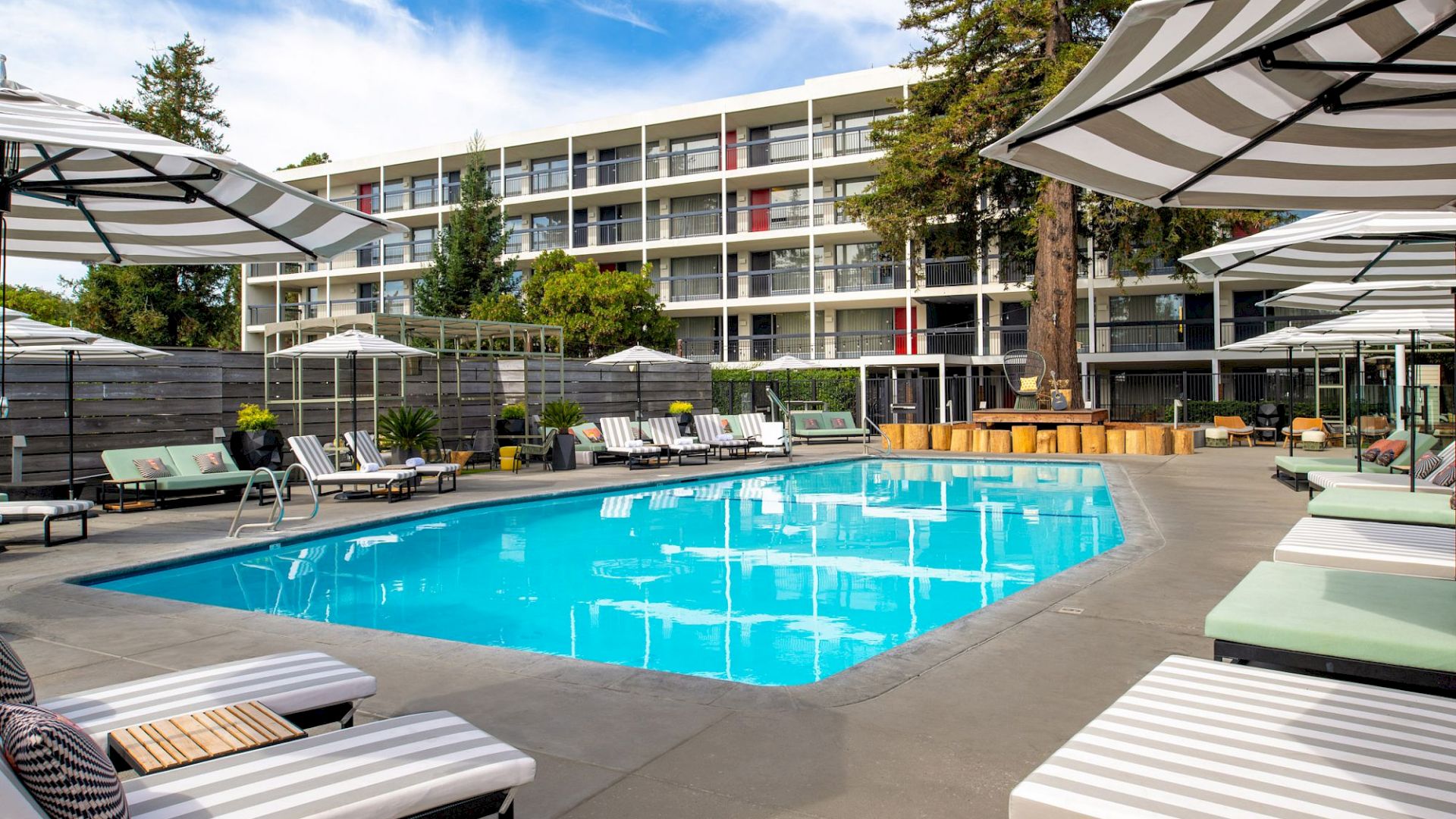 A modern hotel pool area with lounge chairs, umbrellas, and surrounding greenery, set against a backdrop of a multi-story building.