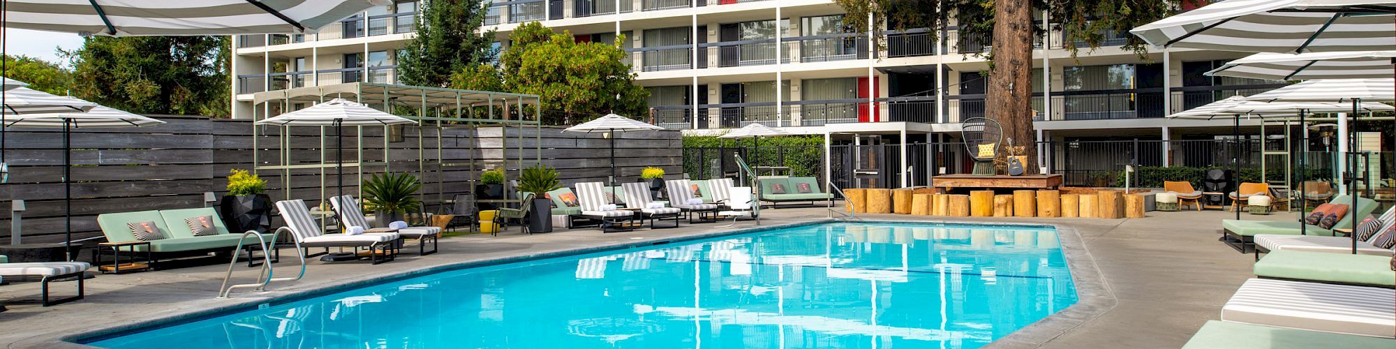 A modern hotel pool area with lounge chairs, umbrellas, and surrounding greenery, set against a backdrop of a multi-story building.