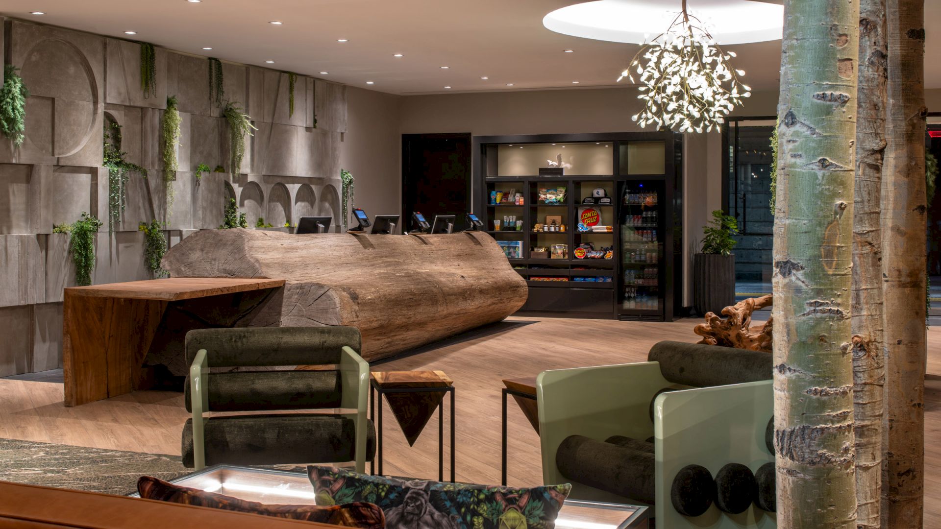 The image shows a stylish hotel lobby with modern decor, featuring a wooden reception desk, seating area, indoor plants, and shelves stocked with items.
