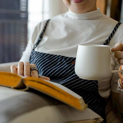 A person is sitting with a book in one hand and a white mug in the other, dressed in a white shirt and striped overalls.