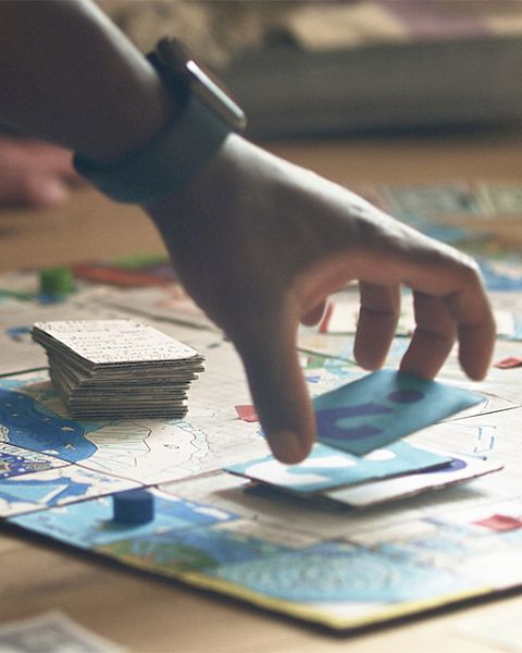 A hand holding a card is about to place it on a board game; other hands are visible, with a stack of cards in the background.