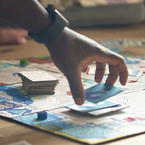 A hand holding a card is about to place it on a board game; other hands are visible, with a stack of cards in the background.